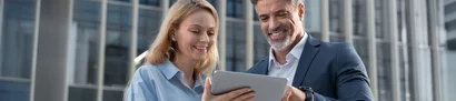 Innovation und Cloud-Technologien entlasten KMU-Führungskräfte Lächelnde Geschäftspersonen schauen sich gemeinsam Inhalte auf einem Tablet vor Bürogebäuden an.
