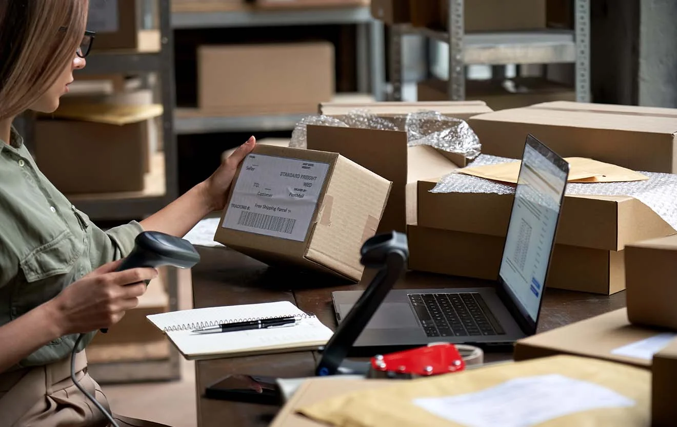 Mobile data capture in distance selling – warehouse management in the ERP system An employee scans a parcel and works on a laptop between boxes.
