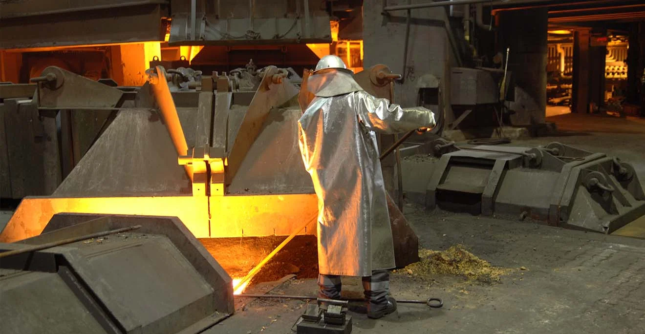 Worker in heat protection clothing operates an open melting furnace with liquid metal in a factory