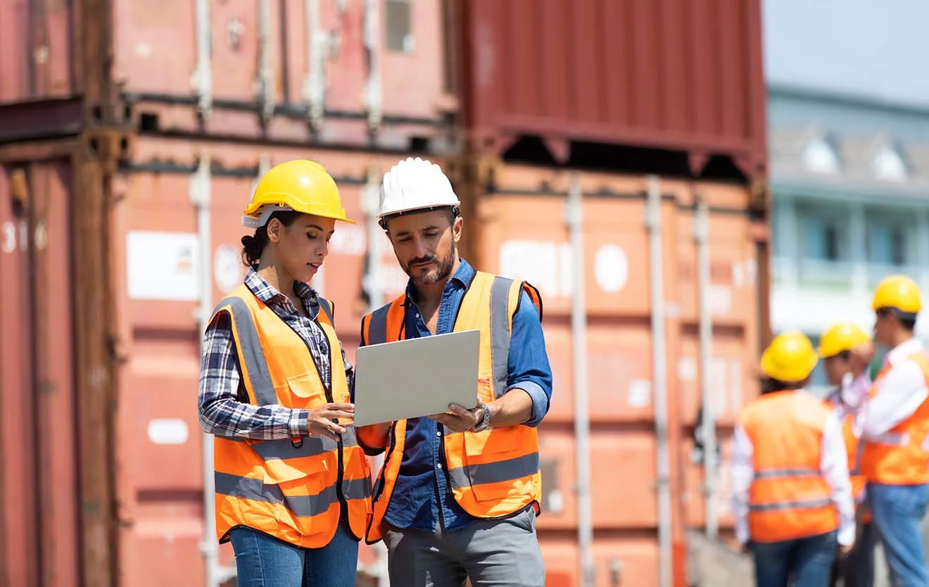 Two employees wearing protective helmets stand in front of containers and work together on a laptop.