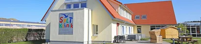 SyltKlinik der Deutschen Kinderkrebsstiftung mit rotem Dach, Terrasse und Strandkorb im Garten.