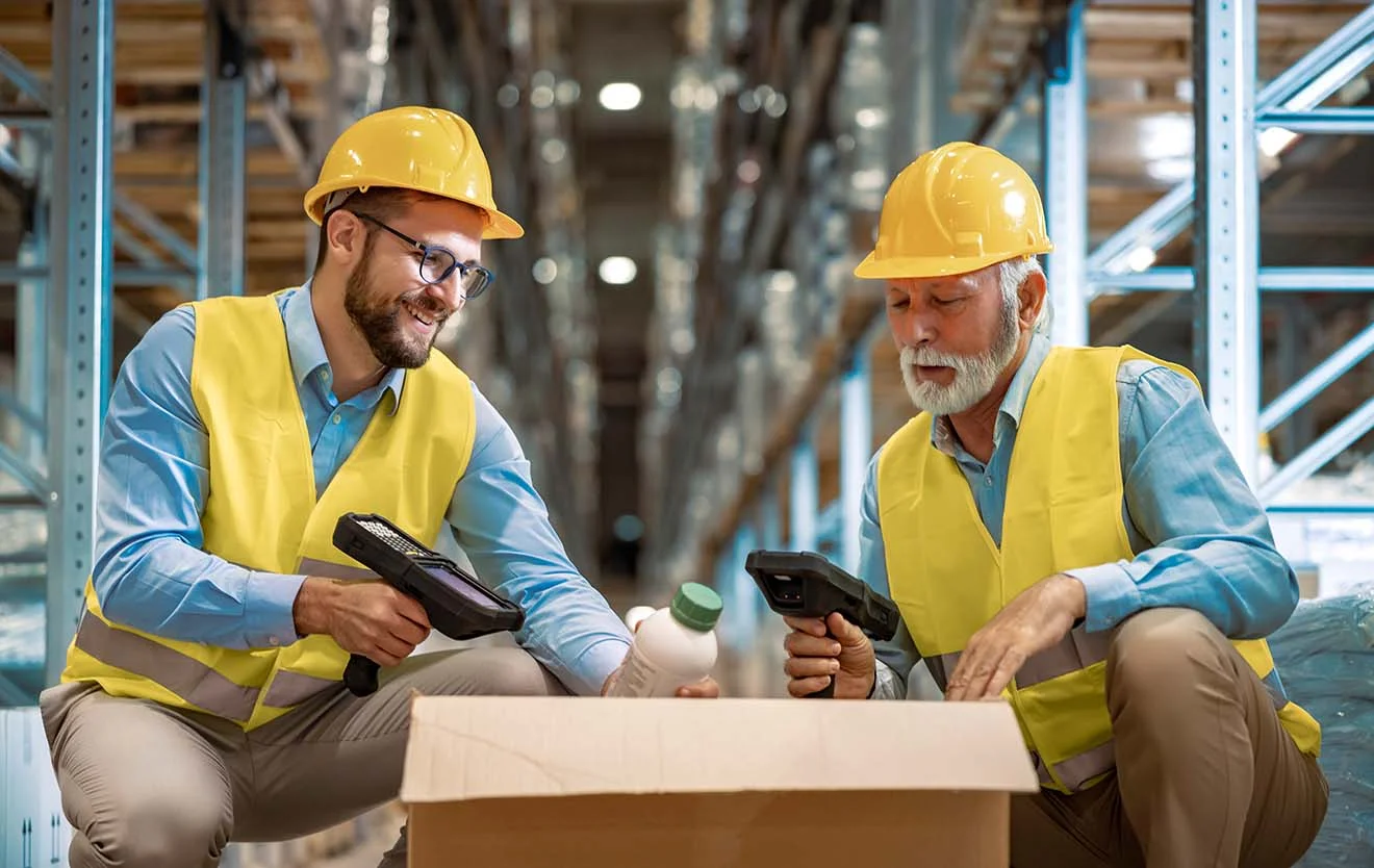 Two warehouse workers in a wholesale business scan products from a box using handheld scanners.