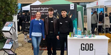 Drei Personen stehen am GOB-Messestand; hinten Plakate, vorne rechts Tisch mit Give-aways.