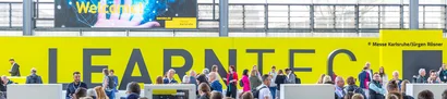 LEARNTEC 2025 in Karlsruhe: Eingang Besucher | 1905 x 425 Pixel Menschenmenge am Eingang der LEARNTEC 2025 in Karlsruhe vor gelber Wand mit Schriftzug
