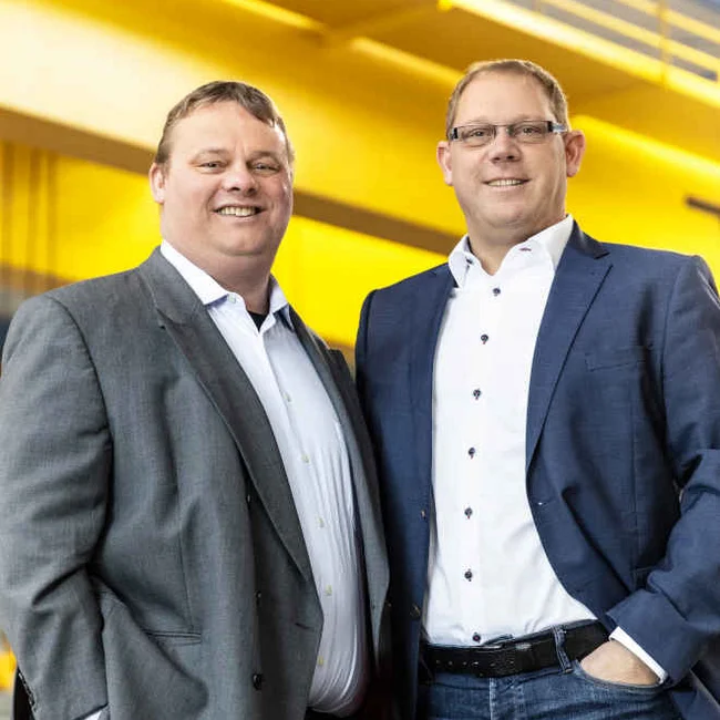 Zwei Männer im Business-Outfit stehen nebeneinander und lächeln; gelber Industriehintergrund.