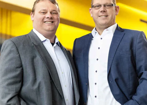 Zwei Männer im Business-Outfit stehen nebeneinander und lächeln; gelber Industriehintergrund.