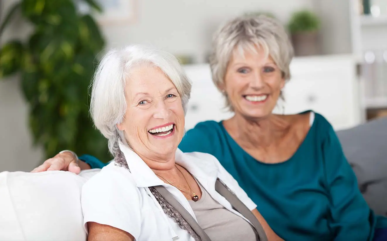 Zwei ältere Frauen lachen gemeinsam auf dem Sofa und genießen ihren sorgenfreien Ruhestand