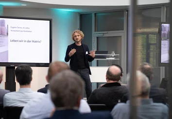 Christian Uhle spricht vor Publikum; Folie „Leben wir in der Matrix?“ auf der Leinwand.