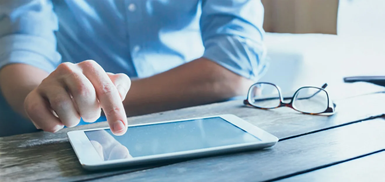 Symbolbild: Hand tippt auf ein Tablet am Tisch, Brille liegt daneben, Hintergrund unscharf.