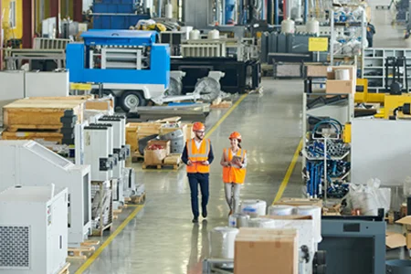 Two people with safety vests walking through an industry hall with machines for variant production.