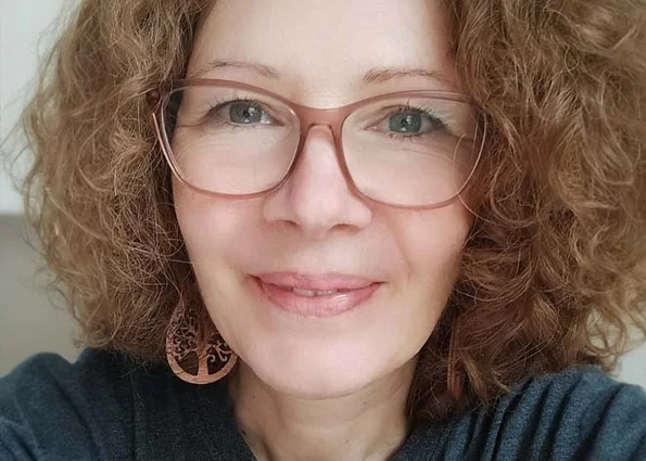 Frau mit lockigen Haaren und großer Brille lächelt in die Kamera vor hellem Hintergrund.