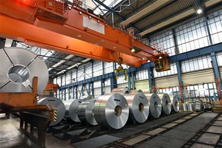 Große Stahl-Coils stehen aufgereiht in einer Produktionshalle mit Krananlage und Lagerbereichen