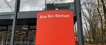 Rote Säule mit Aufschrift Blue Box Bochum vor verglastem Gebäude und Bäumen.