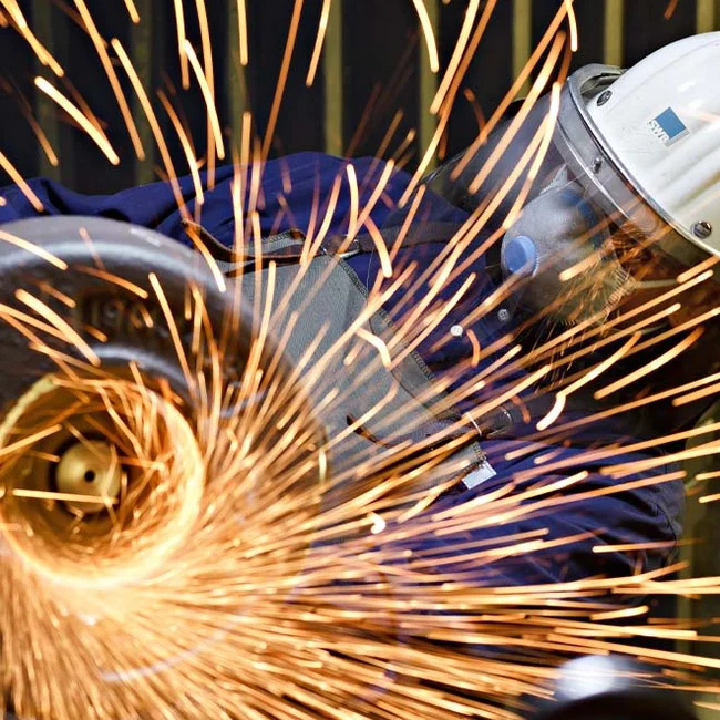 Funken fliegen vom Winkelschleifer; Person mit SWB-Helm und Atemschutz arbeitet am Metall.