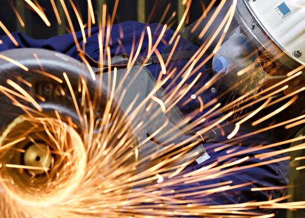 Funken fliegen vom Winkelschleifer; Person mit SWB-Helm und Atemschutz arbeitet am Metall.