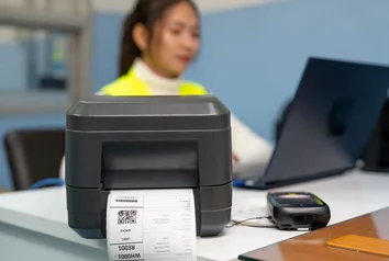 Label printer with shipping label in the foreground, woman with laptop in the background