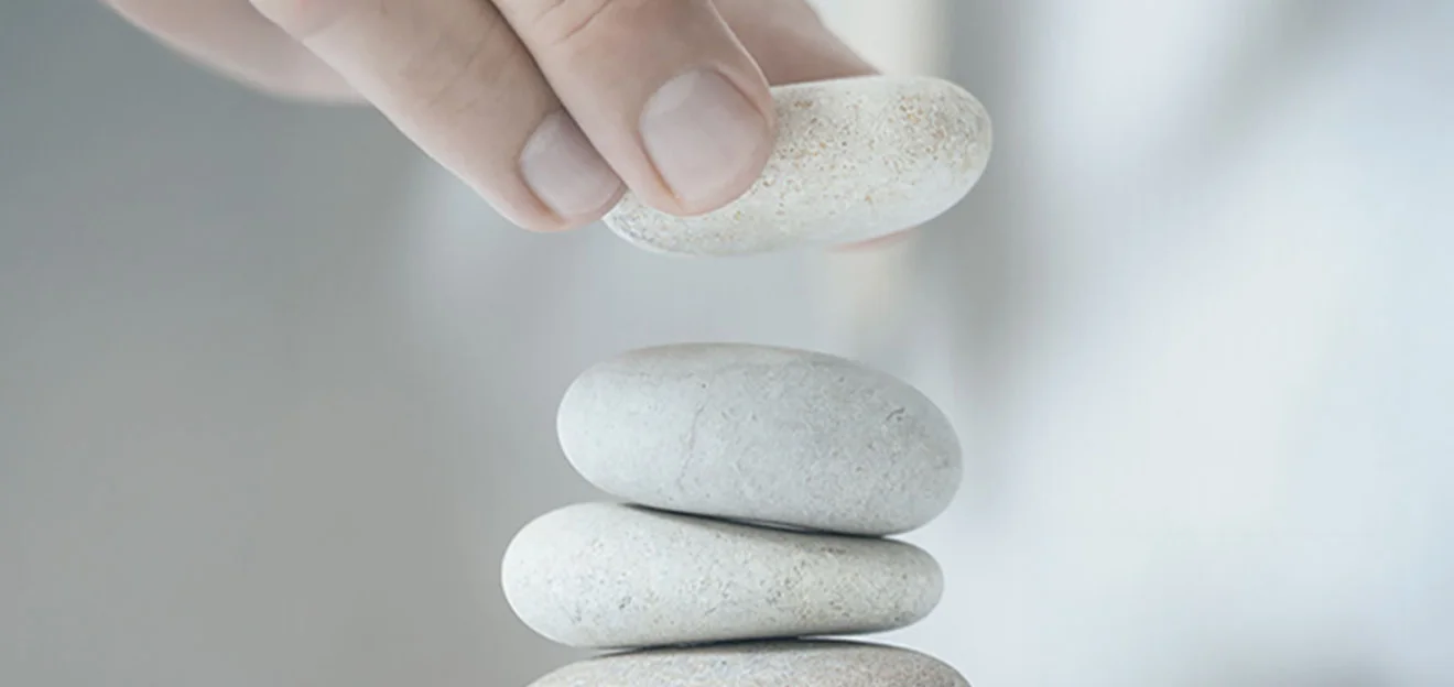 Illustrative image: Hand places a stone on a stack of four pale stones, soft background.