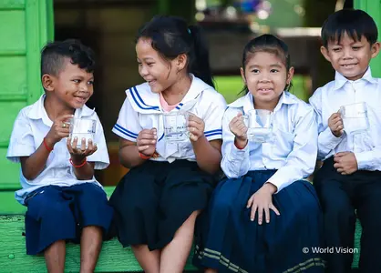 4 lachende Kinder sitzen auf einer grünen Holztreppe mit jeweils einem Glas Wasser in der Hand. Links ist ein volles Trinkwasserbehälter zu sehen.