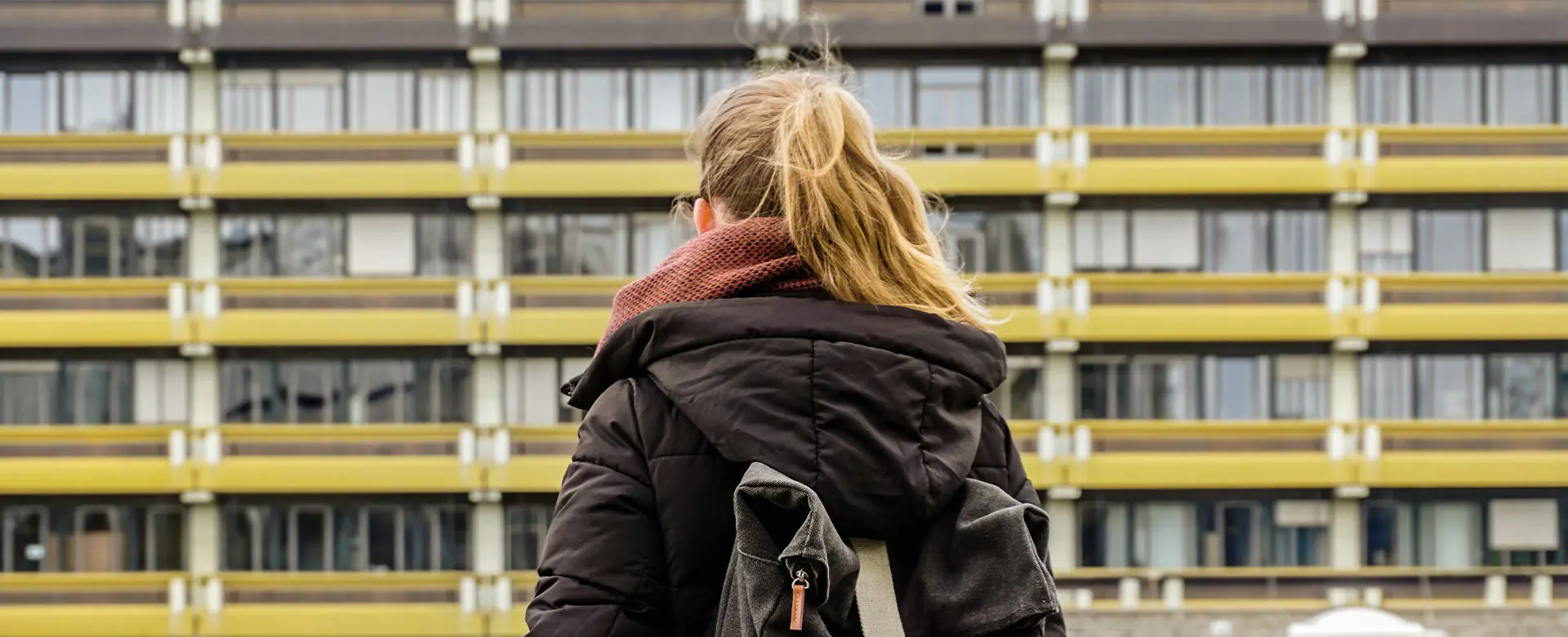 Dunkelblonde Studentin mit Rucksack vor RUB-Gebäude, von hinten aufgenommen.