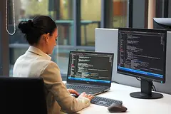 Dunkelhaarige Business-Frau programmiert am Laptop; Codezeilen sind auf beiden Monitoren zu sehen