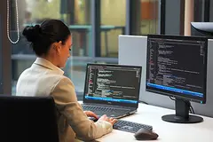 Dark-haired business woman programming on a laptop; lines of code can be seen on both Monitors