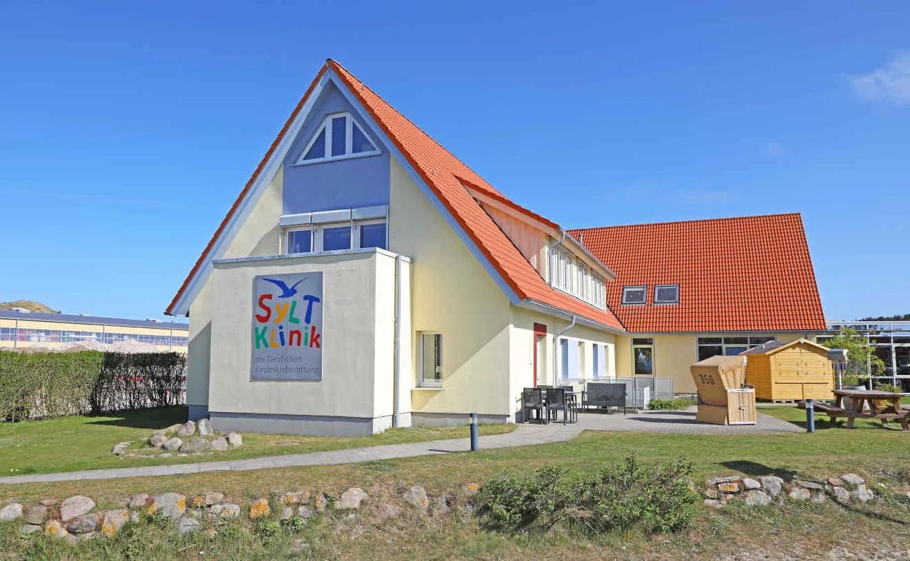Gesamtansicht der gelben SyltKlinik der Deutschen Kinderkrebsstiftung mit Garten und rotem Dach.